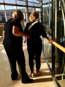 Photo of two women walking, from behind. One woman is helping the other one, who is leaning on a handrail for support