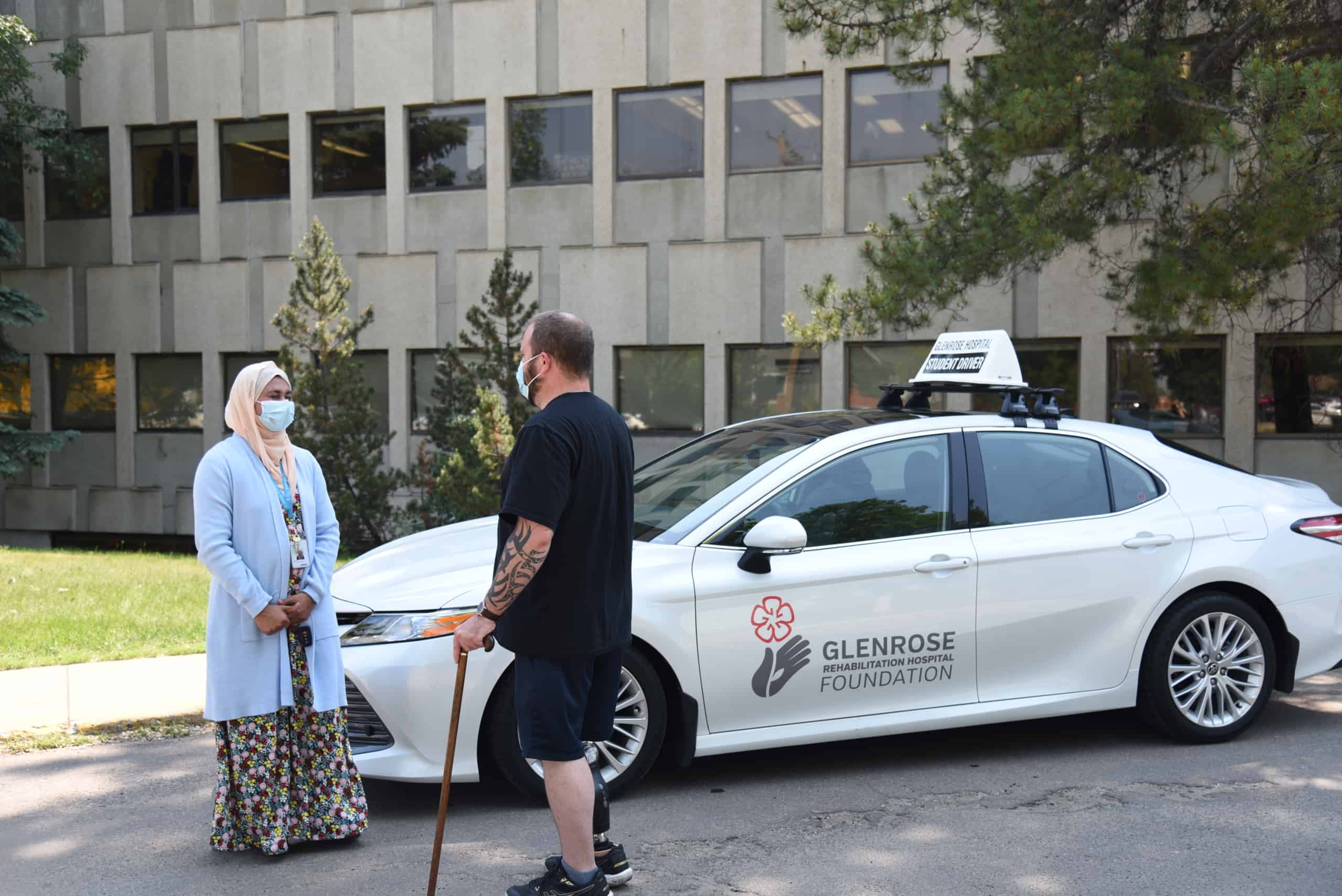 Photo of a man and woman standing outside a white car. They are both wearing masks and are speaking to each other.