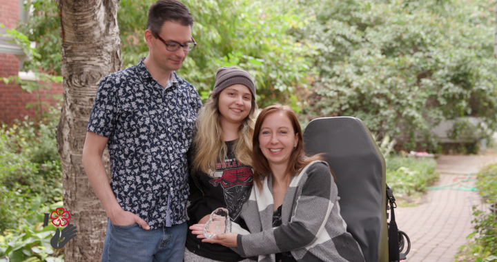 Photo of a woman sitting in a park with her daughter and husband. The seated woman is holding an award.
